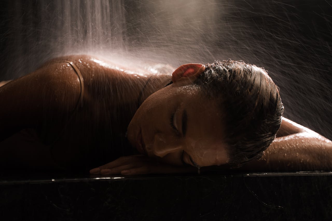 Woman in a spa setting under waterfall, relaxing with eyes closed on a marble surface.